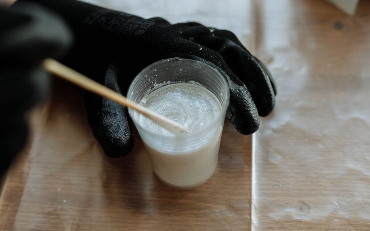 Visão detalhada de mãos com luvas misturando resina em um copo plástico com um agitador em uma superfície de madeira